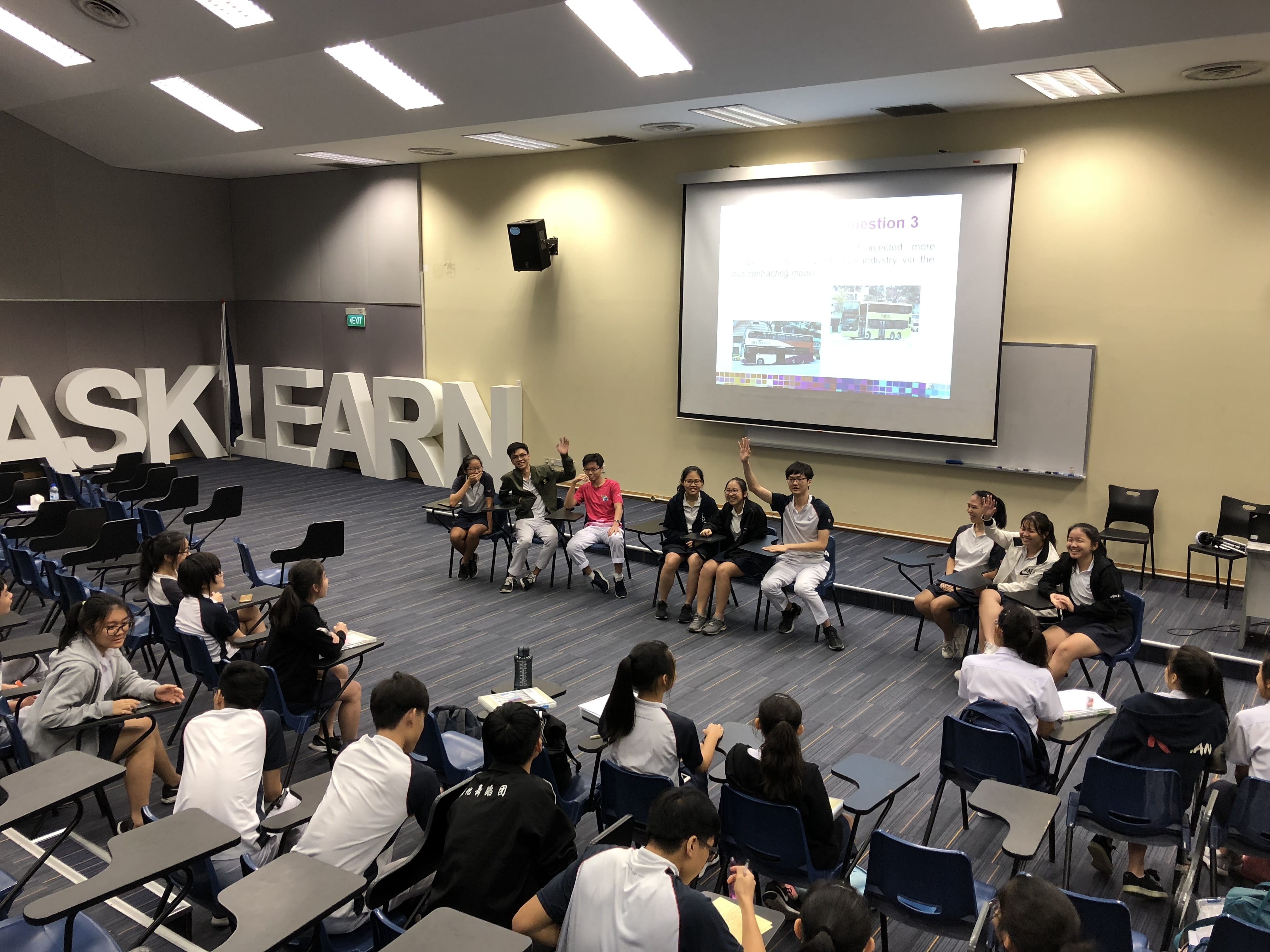 Students in uniforms sit in a circle during a classroom session with a projected slide reading Question 3.